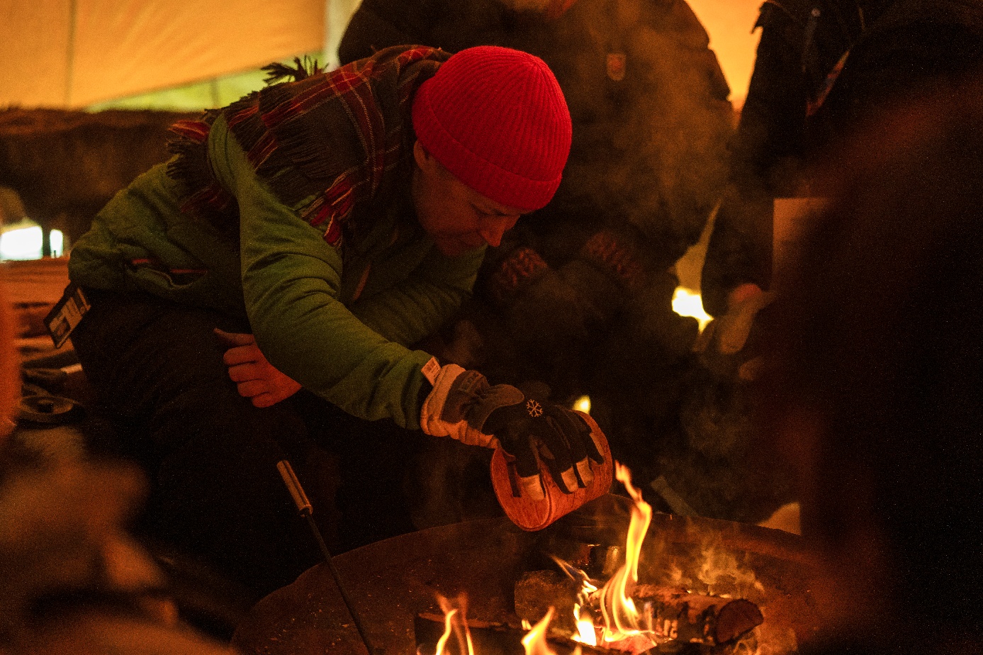A person with a red woolly hat pours coffee ground into a pot on an open fire