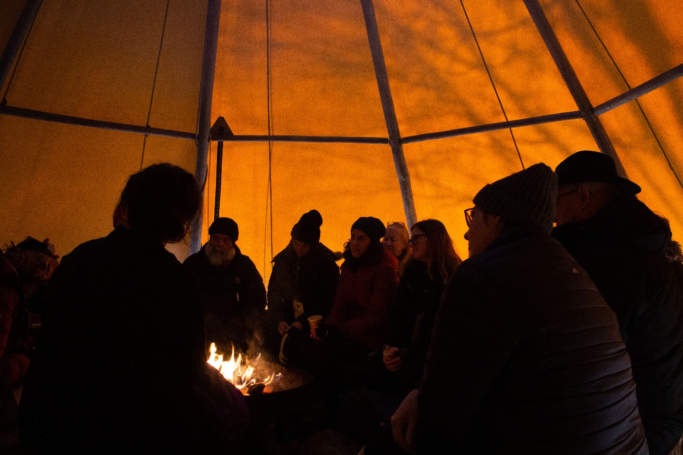 People gathered around a fire in a sunlit tentipi with faint shadows of tree branches on the canvas