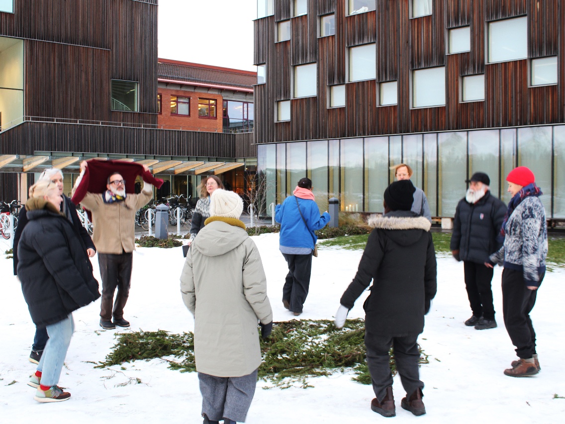 People in a circle jump around a small pile of pine branches outside the Arts Campus of Umeå University, Sweden